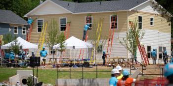 Volunteers working on the site of the 2026 Carter Work Project in the midground. In the background, a multi-family home is being painted by volunteers on ladders, as other volunteers work below. 