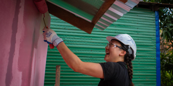 A young woman wearing a hardhat has a big smile on her face as she rolls pink paint on the side of a house.