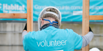 A bright blue shirt that says "volunteer" on the back is worn by a person along with a hardhat and gloves as they hold wood framing together in a build site