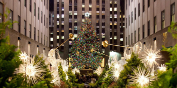 A very large decorated Christmas tree stands in Rockefeller Center plaza in NYC surrounded by decorative lights and angel figures holding golden trumpets.