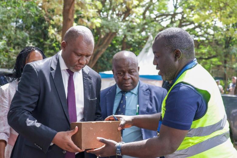 A man in a safety vest hands a building block to a man dressed in a suit