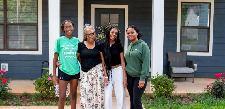 A family of four stands in front of the front door of their home and smiles for the camera.